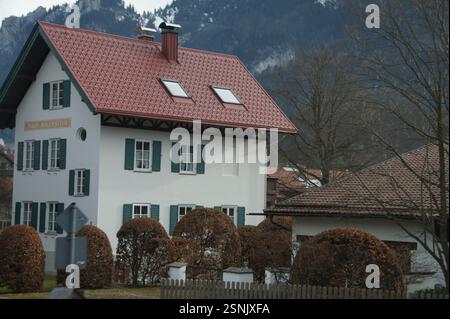 Ein weißes Haus mit rotem Dach und grünen Fensterläden liegt eingebettet zwischen Büschen und Bäumen in Deutschland, München. Die charmante Szene erinnert an die Möglichkeit Stockfoto