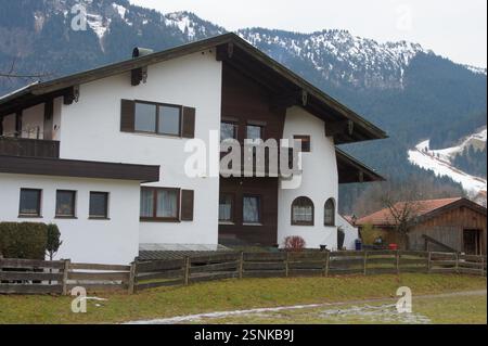 Deutschland, München. Ein weißes Haus mit einem Holzzaun liegt in einer bergigen Region. Die charmante Szene erinnert an ein mögliches Ferienhaus oder eine Vermietung Stockfoto