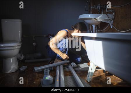 Klempner in Overalls arbeiten unter einem Waschbecken in einem schwach beleuchteten Badezimmer mit sichtbaren Rohren Stockfoto