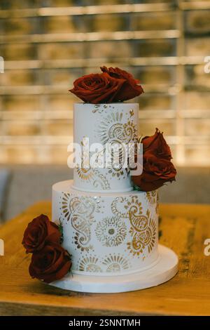 Ein atemberaubender zweistöckiger Kuchen mit roten Rosen und komplizierten Goldmotiven auf einem Holztisch bei einer festlichen Versammlung. Stockfoto