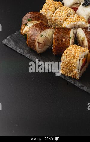 Eine wunderschön angeordnete Platte zeigt eine Vielzahl von Sushi-Rollen, einschließlich einzigartiger Stile und Texturen, vor einem dunklen Schieferhintergrund. Stockfoto