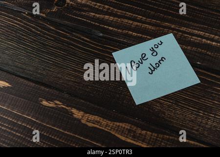 Eine handgeschriebene Notiz auf blauem Klebepapier lautet: I love you Mom ruht auf einem rustikalen Holztisch. Es vermittelt eine herzliche Botschaft der Wertschätzung und des Lichts Stockfoto