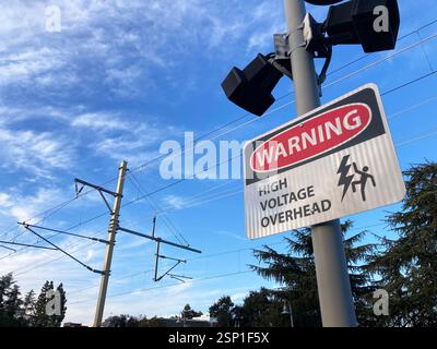 Hochspannungs-Warnschild an einem Stab, das vor elektrischen Kabeln warnt, mit sichtbaren Stromleitungen und einem klaren blauen Himmel im Hintergrund Stockfoto