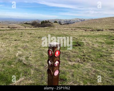 Verwittertes Schild für den Ohlone Wilderness Regional Trail, mit Pfeilen und einer Fahrradikone steht auf einem grasbewachsenen Feld, umgeben von sanften Hügeln - Sa Stockfoto
