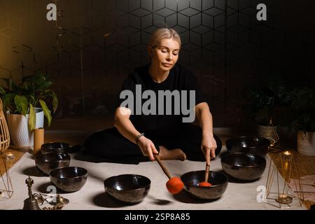 Frau, die während einer friedlichen Abendveranstaltung mit Klangschalen in einem ruhigen, mit Pflanzen und sanfter Beleuchtung dekorierten Innenraum die Klangheilung übt Stockfoto