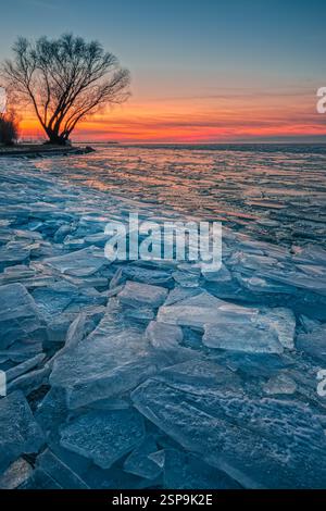 Fonyod, Ungarn - der gefrorene Balaton-See an einem kalten Winternachmittag mit gebrochenem Eis, goldenem farbenfrohem Sonnenuntergang Himmel und Baum im Hintergrund Stockfoto