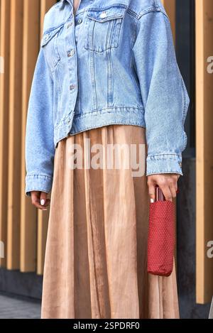 Modische Frau in Jeansjacke und fließendem Rock mit rotem Regenschirm-Accessoire, das ein modernes Stilkonzept repräsentiert Stockfoto