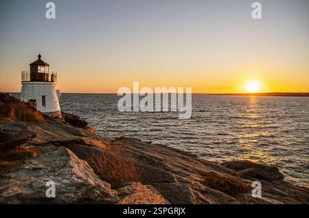 Ein historischer Leuchtturm steht an einer felsigen Küste mit Blick auf das Meer, während die Sonne untergeht, und wirft goldenes Licht über das Wasser in Rhode Island. Stockfoto