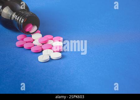 Rosafarbene und weiße Arzneimittelpillen, die aus einer dunklen Flasche auf blauem Hintergrund auslaufen und Gesundheitskonzepte und pharmazeutische Konzepte repräsentieren Stockfoto