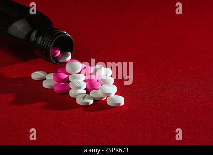 Weiße und rosafarbene Pillen werden aus einer dunklen Flasche auf einem leuchtend roten Hintergrund ausgeschüttet und erzeugen einen starken Kontrast. Stockfoto