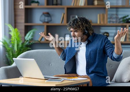 Unzufriedener Mann, der zu Hause auf dem Sofa sitzt, mit Headset und Kopfhörern. Nervöser Freelancer, der per Fernzugriff mit Videoanrufen arbeitet, streitet und schreit. Stockfoto