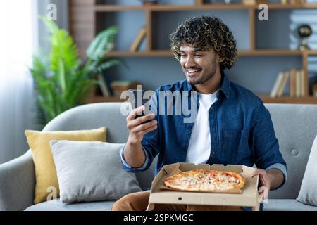 Junger Mann zu Hause mit Pizza und Telefon in der Hand, zufrieden mit der Qualität der Online-Bestellung und der Geschwindigkeit der Lieferung. Glücklicher Kunde bestellte ein Sofa im Wohnzimmer. Stockfoto