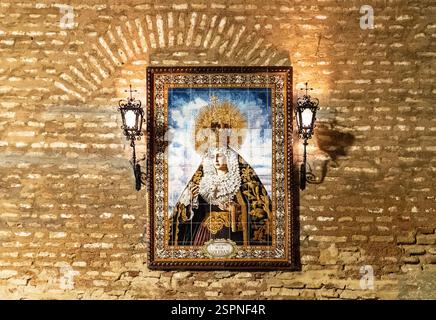 Ein religiöses Kunstwerk, das eine Figur mit Krone und aufwendiger Kleidung darstellt, gerahmt und auf einer strukturierten Ziegelwand montiert. Zwei kunstvolle Wandlampen leuchten Stockfoto
