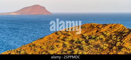 Ein malerischer Blick auf eine felsige Küste mit einer entfernten vulkanischen Insel im Hintergrund. Der Vorderboden weist zerklüftetes Gelände mit karger Vegetation auf, wh Stockfoto