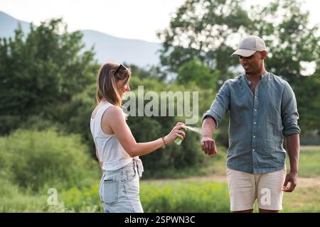 Junges Paar, das während eines Sommerurlaubs Zeckenschutzmittel auf ihre Beine und Arme spritzt Stockfoto