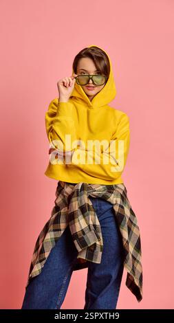 Junge Frau mit selbstbewusstem Ausdruck, verstellbarer Sonnenbrille, gelbem Hoodie und kariertem Hemd vor rosa Studiohintergrund. Stockfoto