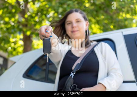 Stolze Geschäftsfrau, die Autoschlüssel vor ihrem neuen Elektroauto hält und vor Zufriedenheit und Begeisterung über ihren letzten Kauf und die Reise lächelt Stockfoto