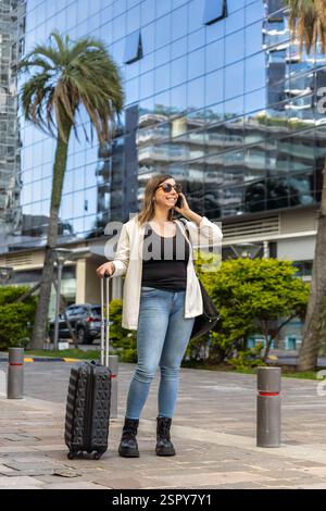Die junge Geschäftsfrau läuft in einer modernen Stadt die Straße entlang, zieht ihr Gepäck, spricht mit ihrem Smartphone und genießt den Komfort eines Mobiltelefons Stockfoto