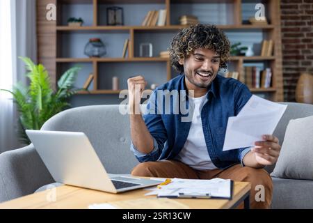 Erfolgreicher zufriedener Mann, der zu Hause mit Dokumenten und Rechnungen arbeitet, im Wohnzimmer auf dem Sofa sitzt, Laptop in Papierkram verwendet, große Erfolge feiert und die Hand hochhält. Stockfoto