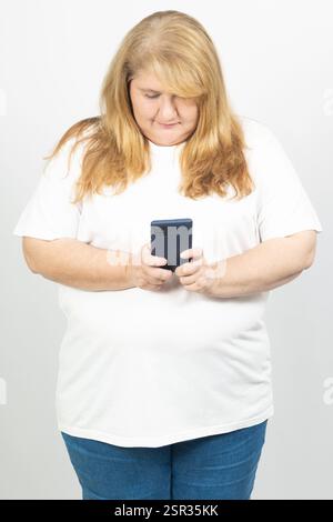 Porträt einer blonden Frau in Übergröße, die ein Smartphone hält. Sie steht entspannt in einem weißen T-Shirt und einer blauen Jeans. Stockfoto