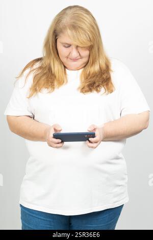 Porträt einer großen blonden Frau, die ein Smartphone horizontal hält. Sie konzentriert sich auf das Gerät und trägt ein lässiges weißes T-Shirt und Jeans. Stockfoto