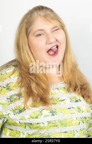 Porträt einer Reifen Frau in Übergröße mit blonden Haaren und grünen Augen, die spielerisch zuzwinkernd und lächelnd wirkt. Ihr ausdrucksstarkes Gesicht und ihr lebhaftes Oberteil Stockfoto
