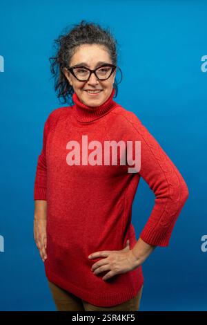 Porträt einer fröhlichen, Reifen Frau mit Brille und rotem Rollkragenpullover, die selbstbewusst mit Händen auf den Hüften vor einem leuchtend blauen Backdro posiert Stockfoto