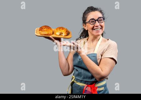 Lächelnde Bäckerin, die ein Holztablett mit zwei köstlichen frisch gebackenen Zimtrollen hält und zeigt Stockfoto