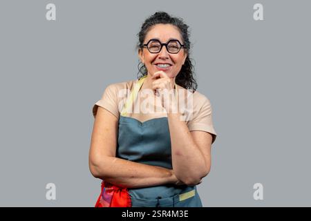 Porträt einer fröhlichen reifen Frau mit Schürze, die lächelnd mit der Hand auf dem Kinn denkt, vor grauem Hintergrund Stockfoto