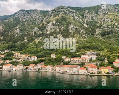 Bucht von Kotor, Montenegro - 2. Juli 2024: Muo Village. Küstenlinie. Autos parken entlang der Wasserlinie mit roten Gebäuden. Grüne Bäume auf grauem felsigem Moun Stockfoto