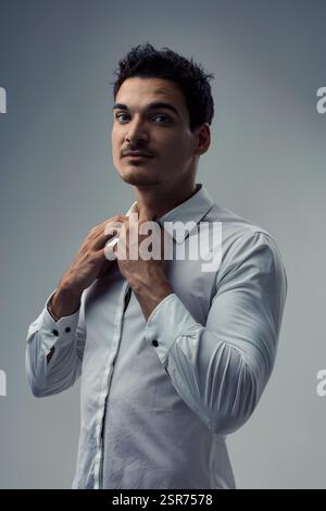 Studio-Porträt eines selbstbewussten und stilvollen jungen Mannes, der seinen weißen Hemdkragen vor einem grauen Hintergrund anpasst und Eleganz und Charme ausstrahlt Stockfoto