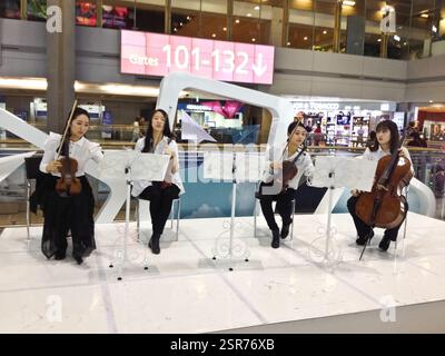 Die Geiger sind in formeller Kleidung gekleidet und spielen ihre Instrumente. Der Flughafen ist voll mit Passagieren und Personal. Violinmusik ist oft pl Stockfoto