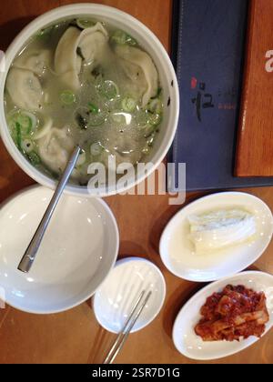 Koreanische Knödelsuppe mit Kimchi und eingelegtem Kohl. Kleine weiße Schüsseln mit Essstäbchen, Löffel und Knödelsuppe in einer großen Schüssel. Kimchi und eingelegt Stockfoto
