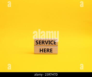 Symbol „Service here“. Concept Words Service hier auf Holzblöcken. Schöner gelber Hintergrund. Business and Service Here Konzept. Kopierbereich. Stockfoto