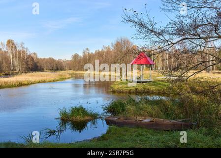 Ein ruhiger Blick auf einen Fluss tagsüber, mit einem alten, verlassenen Holzboot am grasbewachsenen Fluss und einem malerischen roten Pavillon in der Nähe Stockfoto