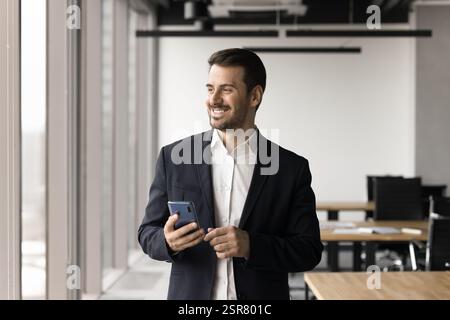 Lächelnder, gutaussehender Geschäftsmann im Anzug, der ein Smartphone hält Stockfoto