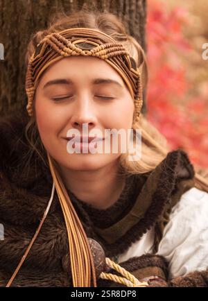 Das Porträt eines Kriegermädchens in historischem Kostüm sitzt neben einem Baum im Herbstwald Stockfoto