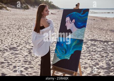 Die junge Malerin steht am Sandstrand und beobachtet ihre lebendigen Kunstwerke auf der Staffelei. Malerei zeigt abstrakte ozeanische und kosmische Themen, Ref Stockfoto