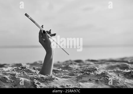 Die weibliche Hand taucht anmutig aus dem Sand auf und hält einen Vintage-Zigarettenhalter. Das monochromatische Bild am ruhigen Strand weckt Neugier und E Stockfoto