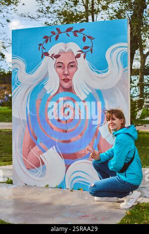 Junge Künstlerin mit braunen Haaren und blauer Jacke kniet neben einem lebendigen Wandgemälde mit einer Frau mit fließenden Haaren und Spiralmustern, die unter der Sonne steht Stockfoto