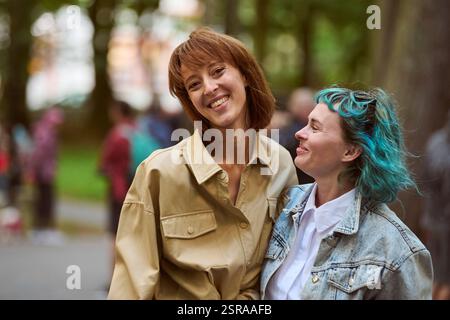 Junge Frauen stehen dicht im Park. Eine mit braunem Haar, beige Jacke, andere mit blauem Haar, Jeansjacke. Beide lächeln. Unschärfe von Bäumen, Menschen Stockfoto
