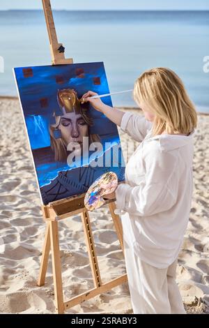 Junge Künstlerin, die mystische Porträts auf Leinwand am ruhigen Strand malte. Das Kunstwerk zeigt ein surreales weibliches Gesicht, umgeben von abstrakten Elementen, die evozieren Stockfoto