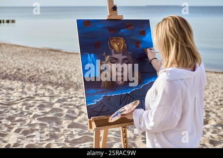 Junge Künstlerin, die mystische Porträts auf Leinwand am ruhigen Strand malte. Das Kunstwerk zeigt ein surreales weibliches Gesicht, umgeben von abstrakten Elementen, die evozieren Stockfoto