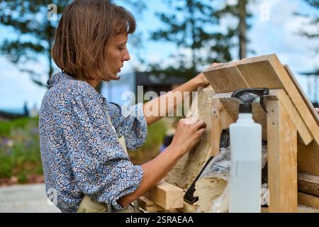 Junge Erwachsene Frau mit braunem Haar schafft Tonreliefkunst draußen. Mit gemustertem Hemd, fokussiertem Ausdruck. Bäume und Holzstruktur sichtbar Stockfoto