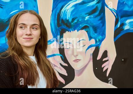 Junge Erwachsene Frau mit braunen Haaren steht neben farbenfroher Leinwand. Das Wandbild zeigt ein stilisiertes Gesicht mit geschlossenen Augen und fließendem blauem Haar. Tagesbeleuchtung A Stockfoto