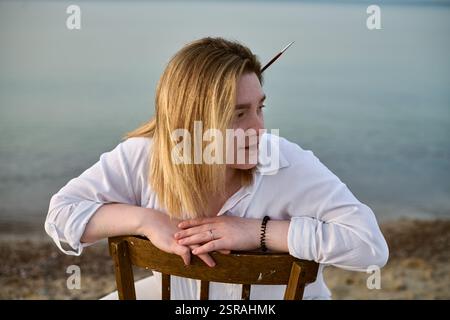 Junge Frau in weißem Hemd entspannt sich auf Holzstuhl am ruhigen Meer. Ihr nachdenklicher Blick und ihre lässige Pose spiegeln Ruhe und Selbstsicht in Ruhe wider Stockfoto