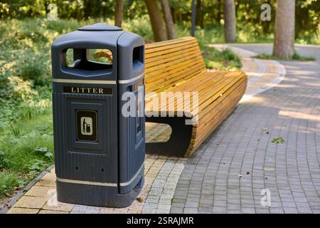 Parkbank aus Holz neben Abfalleimer auf sonnendurchflutetem Steinweg in ruhiger Waldlage, einladende Erholung und achtsame Entsorgung. Friedliches Ambiente in üppiger Umgebung Stockfoto
