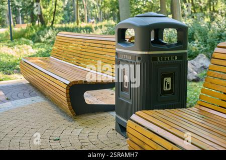 Parkbank aus Holz neben Abfalleimer auf sonnendurchflutetem Steinweg in ruhiger Waldlage, einladende Erholung und achtsame Entsorgung. Friedliches Ambiente in üppiger Umgebung Stockfoto