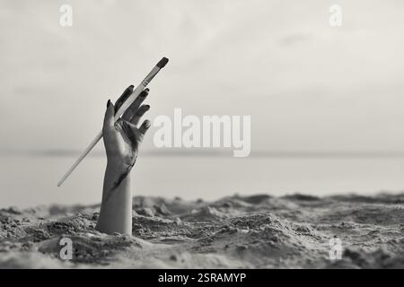 Die weibliche Hand taucht anmutig aus dem Sand auf und hält einen Vintage-Zigarettenhalter. Das monochromatische Bild am ruhigen Strand weckt Neugier und E Stockfoto
