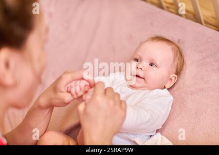 Weißes Mädchen mit hellbraunem Haar, das glücklich auf einer rosa Decke liegt. Erwachsene alleinerziehende Mutter hält sanft die Babyhände. Sanfte Beleuchtung schafft Krieg Stockfoto